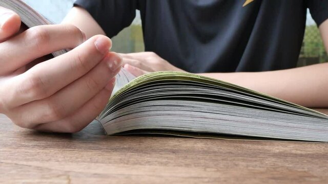 A young teenage boy is reading a comic book at home or library. Close up book on table and fingers hand of man student in black t-shirt sitting in a room with natural daylight from window.   