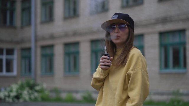 Pretty young hipster woman in black cap yellow hoodie vape ecig. Girl vape popular ecig gadget, vaping device.Happy brunette vaper girl with e-cig. Portrait of smoker female model with electronic