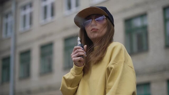 Pretty young hipster woman in black cap yellow hoodie vape ecig. Girl vape popular ecig gadget, vaping device.Happy brunette vaper girl with e-cig. Portrait of smoker female model with electronic