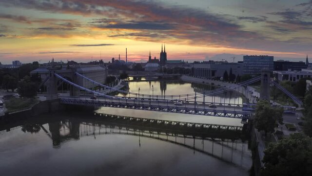 Flyingover Oder river to Grunwaldzki Bridge in Wrocław. Passing bye rushing traffic on the bridge. Illuminated historic buildings and bridges. Beautiful sky, light reflections on blurred water