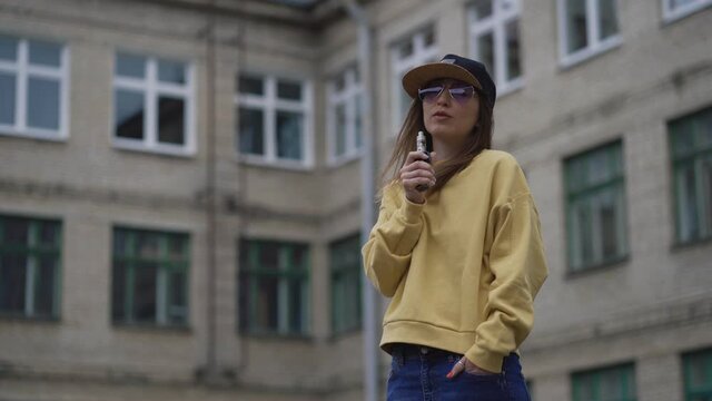 Pretty young hipster woman in black cap yellow hoodie vape ecig. Girl vape popular ecig gadget, vaping device.Happy brunette vaper girl with e-cig. Portrait of smoker female model with electronic
