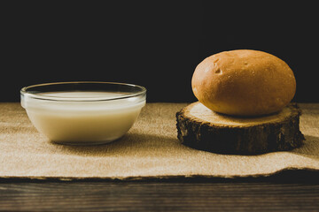 A hamburger bun lies on a section of a tree trunk on a textured fabric, next to a transparent Cup of milk, the background is black, close-up.