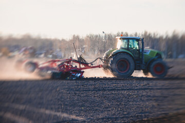 Fototapeta premium Tractor with a disc harrow system harrows the cultivated farm field, process of harrowing and preparing the soil, tractor seeding crops at field on sunset, agriculture concept, harrow machine at work