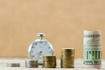 Several stacks of coins with dollar bill and clock in the background out of focus
