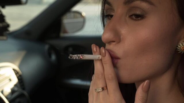Close-up of an oriental-looking girl who sits in the car and cinematically smokes a cigarette, she looks into the distance, inhales and exhales white smoke.