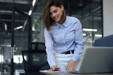 Attractive cheerful business woman checking paper documents in office, working on laptop.