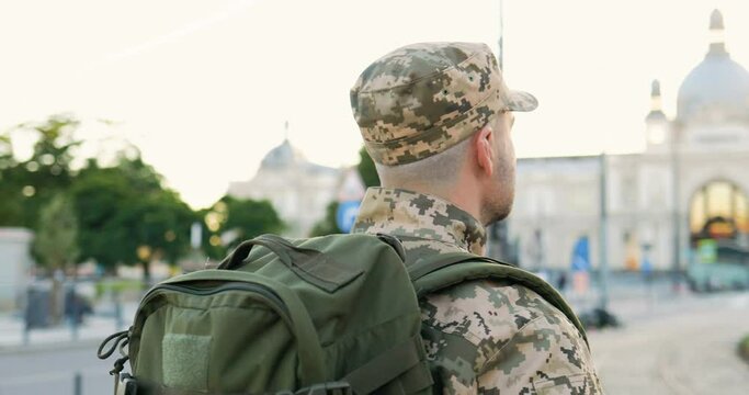 Rear of Caucasian young male soldier in uniform, hat and with backpack walking outdoor. Back view on militarian officer strolling the street in military clothes. Going to army.