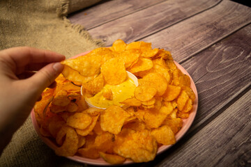 The girl takes a chip from a round dish with potato chips and a saucepan with cheese sauce in the center of the plate. Close up.