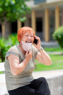 Mature Asian Woman Sitting On A Bench Pulling Down Her Face Mask While Talking On Her Phone.