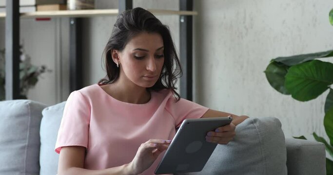 Indian ethnicity young woman resting at home spend free time sitting on couch with tablet electronic portable modern device. On-line internet services and distance communication satisfied user concept