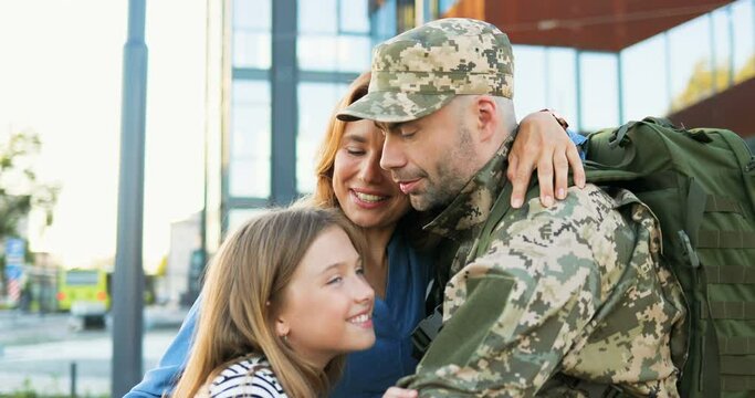 Happy young Caucasian father officer meeting and hugging cute small kid and wife at street. Handsome male soldier in uniform coming back from army and meeting woman with daughter War service returning