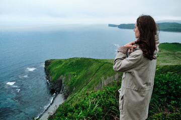 back view of a girl standing in the grass at the edge of a cliff with sky and sea background. Journey and outdoor concept.