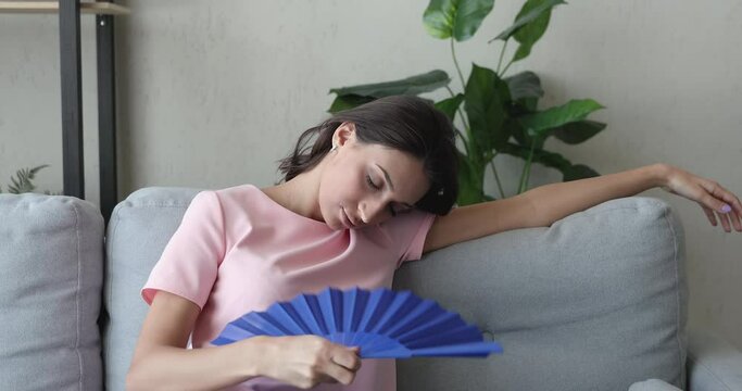 Sweaty young middle eastern ethnicity woman suffering from heatstroke holding handheld blue fan waving cooling herself lean on couch in living room, summer hot weather, need air conditioner appliance