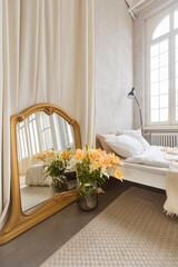 Bouquet of flowers and mirror placed near curtain and comfortable bed in elegant bedroom at home