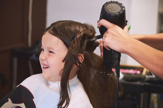 A Little Girl In A Barbershop. Children's Haircut, Hair Drying, Combing And Styling. The Child Is Happy And Contented. Barber Station Wagon.