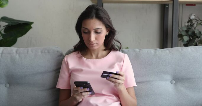 Mixed race Indian woman sit on sofa holds credit card use phone having problems, insufficient funds lack of money, unable withdrawal transaction impossible, insecure online payment, scam fraud concept