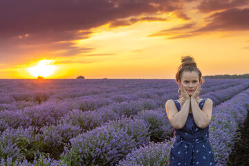 Girl staying near blooming blossoming beautiful landscape of violet purple lavender flowers on field with summer sunset and orange sky, Bulgaria. Essential oils production concept.