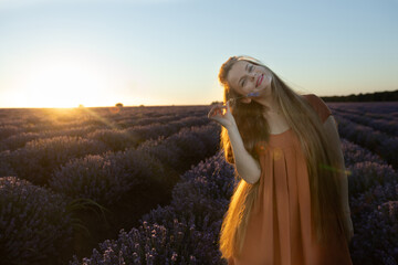 Girl with long hair holds a lavender flower in blooming blossoming beautiful landscape of violet purple lavender field with summer sunset and orange sky, Bulgaria. Happy woman smiles. Beauty concept.