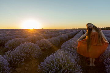 Girl with long hairs staying near blooming blossoming beautiful landscape of violet purple lavender flowers on field with summer sunset, Woman holding her orange dress. Beauty concept.