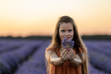 Smiling girl with long hair holds a lavender bouquet in blooming blossoming beautiful landscape of violet purple lavender field with summer sunset and orange sky. Selective focus. Beauty concept.