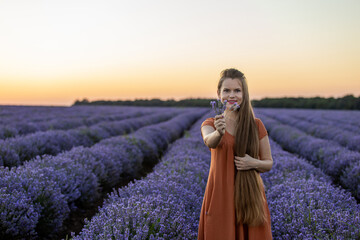 Smiling happy girl in orange dress with long hair holds a lavender bouquet in blooming blossoming beautiful landscape of violet purple lilac lavender field with sunset and orange sky. Summer concept.