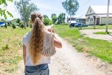A girl with long curly hair, braided in a ponytail, walks to the store along a village road, rear view. Eco-friendly reusable mesh bag, handmade, macrame. Rural landscape. Eco concept without waste.