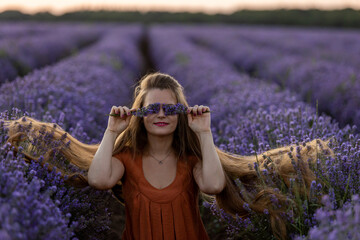 Smiling pretty romantic girl with long hair and in orange dress holds a bunch of lavender flowers and closes her eyes with them. Girl sits in a violet lavender field at sunset. Summer travel concept.
