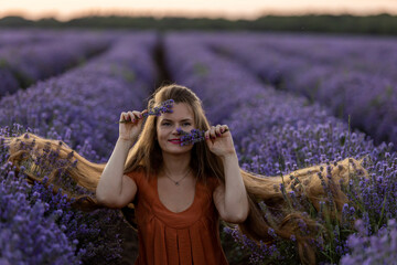Smiling beautiful girl with long hair and in orange dress holds a bunch of lavender flowers and enjoys the fragrance. Girl sits in an incredible violet lavender field at sunset. Summer travel concept.