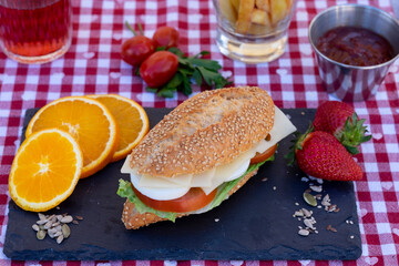 Above view of a black stone cutting board with sesame seed bun with vegetables, eggs and cheese. Checkered tablecloth, red wine glass