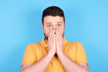 Brunette man scared, holds his hands ove mouth. Isolated on blue background.