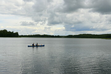 Fototapeta na wymiar Two people swimming in a kayak or small boat traversing a vast yet shallow river or lake with some dense forest or moor visible in the background and a stormy, moody sky above the horizon