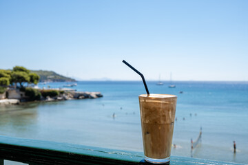 Glass of iced coffee with a straw on a wooden fence with blue sea at the background.