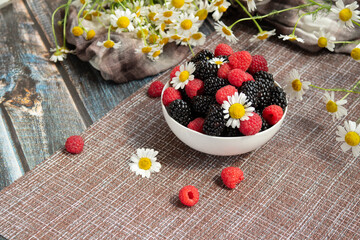 blackberries and raspberries in a white container on a wooden background. Textile. Wild flowers, daisies. Horizontal position of the photo