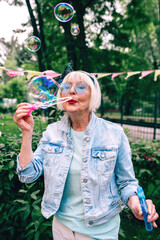 senior (old) stylish woman with gray hair and in blue glasses and jeans jacket blowing bubbles outdoors. Holidays, party, anti age, fun concept