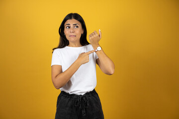 Young beautiful woman standing over isolated yellow background surprised and pointing his watch...