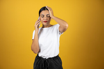 Young beautiful woman talking on the phone with a worried expression on yellow background. Wearing white t-shirt, black shorts and red lipstick