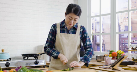 young elegant housewife in apron holding sharp knife slicing cucumber on wooden board at kitchen island. beautiful lady prepare salad. female vegetarian making healthy vegan food at home in city.