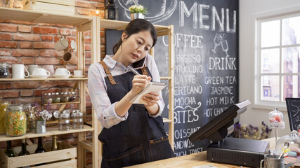 Young Asian woman barista wear apron talking and receive order from customer on cellphone at coffee shop. Concept of cafe store small business. female bartender writing note while listening client.