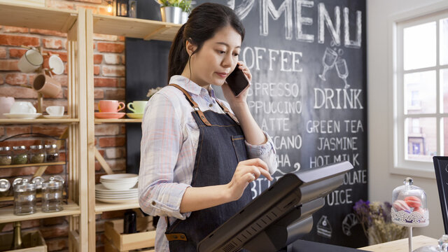 coffee shop assistant taking order on mobile phone in cafe restaurant. young girl waitress in apron standing in counter using point of sale terminal in store while listening to customer talking