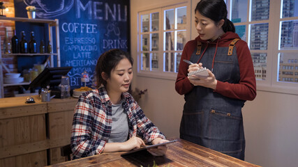 asian japanese waitress taking order on notebook in cafe restaurant. young girl customer sitting alone in night club and talking to bartender. female client holding digital tablet looking at menu.