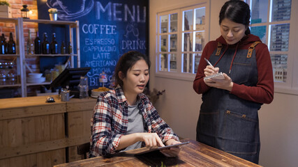 japanese female bartender in apron taking order writing on notepad at table in pub in night. elegant young girl customer talking while looking menu on digital tablet. bar service lifestyle concept.