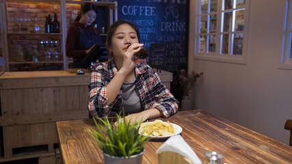 Sad and troubled asian woman at bar alone in night. Lonely depressed female drinking bottle of beer in pub with chips on table. bartender working in background counter using digital tablet in pub.