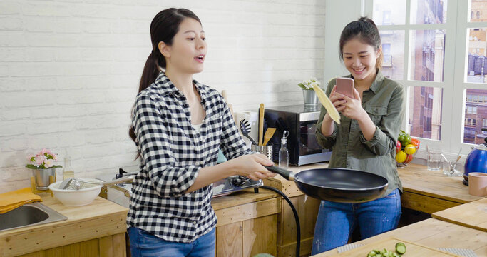 Two female friends cooking and taking photo with cellphone in kitchen. young girl flipping pancake with frying pan while roommate using mobile phone recording video of making process and share online