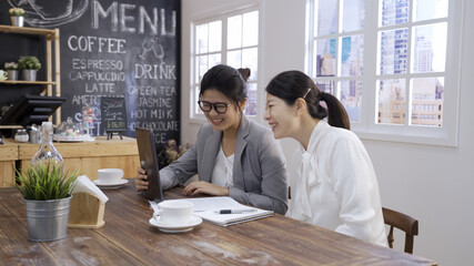 two smiling trendy business women sharing tablet while having business meeting in cafe sitting at table with cups of coffee. young office ladies colleagues using digital pad in coffeehouse indoors.