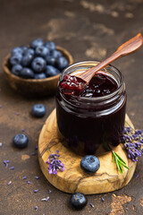 Glass jar and spoon with delicious blueberry jam with fresh berries, lavender and rosemary on dark brown table. Healthy food for breakfast.