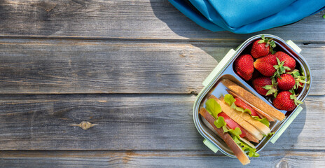 Lunchbox with sandwiches, fresh strawberries, blue chinldren's backpack on wooden bench background. Back to school after quarantine. Lunch break, box with fresh berries. Mom's care for kid's health