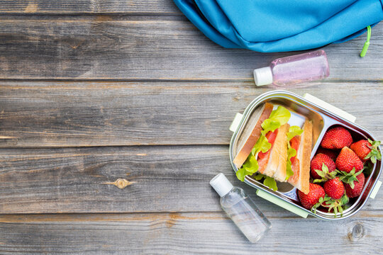 Lunchbox with sandwiches,fresh strawberries,two bottles of sanitizer and blue backpack on wooden background. Back to school after quarantine. Lunch break, box with safety precautions after coronavirus