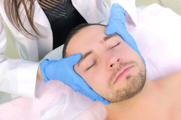 Fototapeta na wymiar Cosmetologist in gloves is applying cream on client's man face massaging skin, top view. Beautician making beauty facial skincare procedure in clinic, portrait of guy patient. Beauty industry concept.