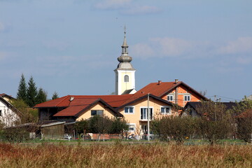 Renovated old church with new facade and metal roof rising above new and unfinished suburban family houses surrounded with dry uncut grass and trees on cloudy blue sky background