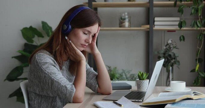 Pensive sad woman in headphones sit at desk in front of laptop thinking, has difficulties, search problem solution, task making, feeling boredom or tired after long studies or exam preparation concept
